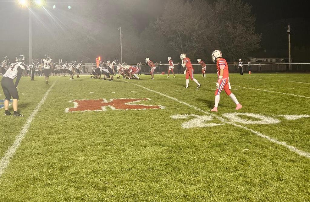 Football players in red uniforms prepared for a play on the field, with a scoreboard in the background and a clear night sky.