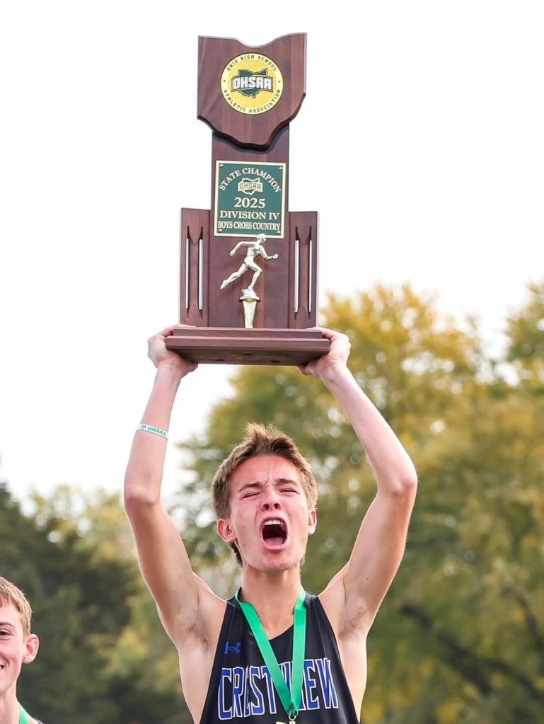 A young male athlete celebrating while lifting the state championship trophy for Division IV Boys Cross Country in 2025, wearing a medal and showing excitement.