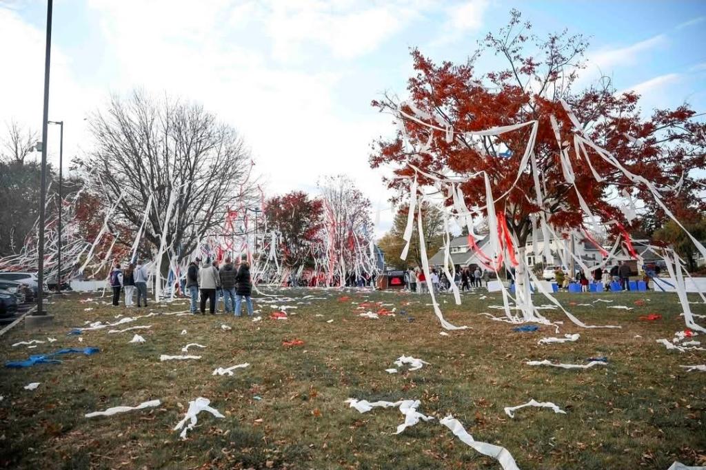 A festive scene in a park featuring trees decorated with toilet paper streamers, surrounded by a group of people celebrating.
