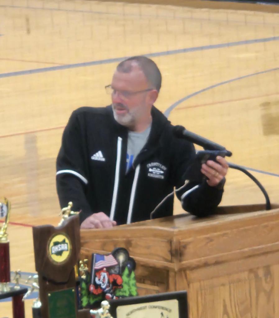 Head coach Randy Grandstaff speaking at a podium in a gymnasium during a pep assembly, with various trophies and awards visible in the foreground.