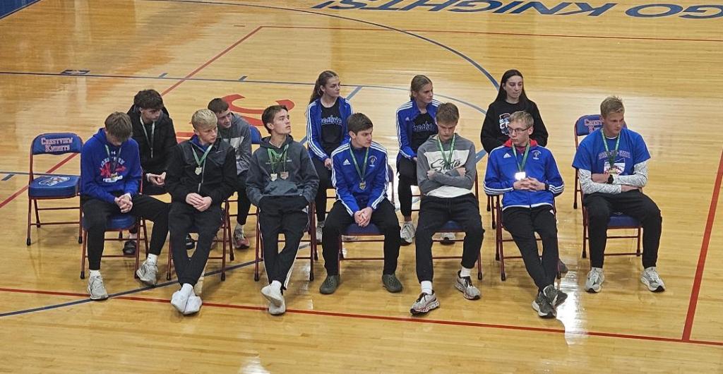 Members of the Crestview Knights cross country team seated in a gymnasium, wearing their medals and team attire, reflecting on their recent state championship victory.