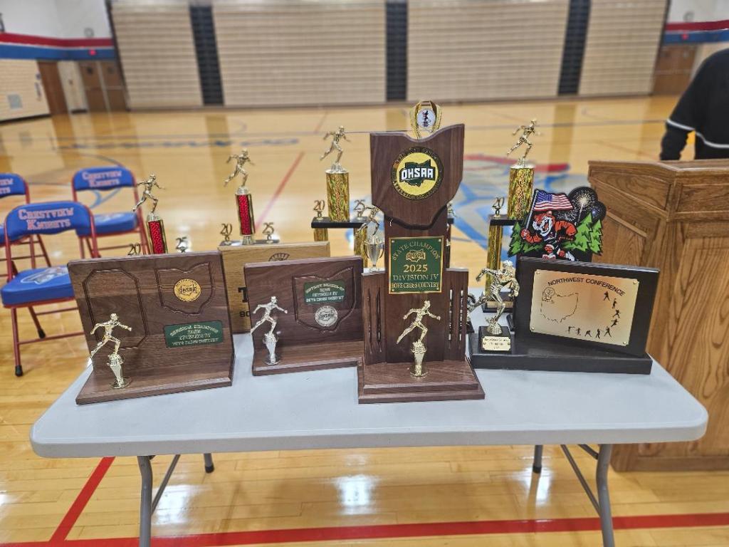A display of trophies and awards celebrating the Crestview Knights' achievements in cross country, with the state championship trophy prominently featured on a table in a gymnasium.