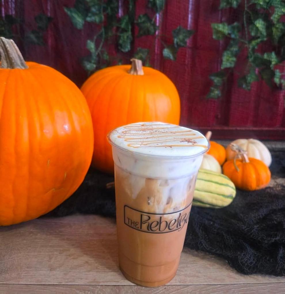 A close-up of a seasonal drink from The Rebel Bean, topped with foam and caramel, set against a backdrop of orange pumpkins and gourds, creating a cozy autumn atmosphere.
