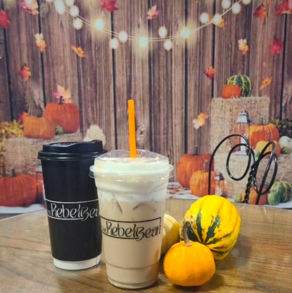 Two coffee cups from The Rebel Bean, one hot and one iced, placed on a wooden table alongside small pumpkins and gourds, with a festive autumn-themed backdrop.