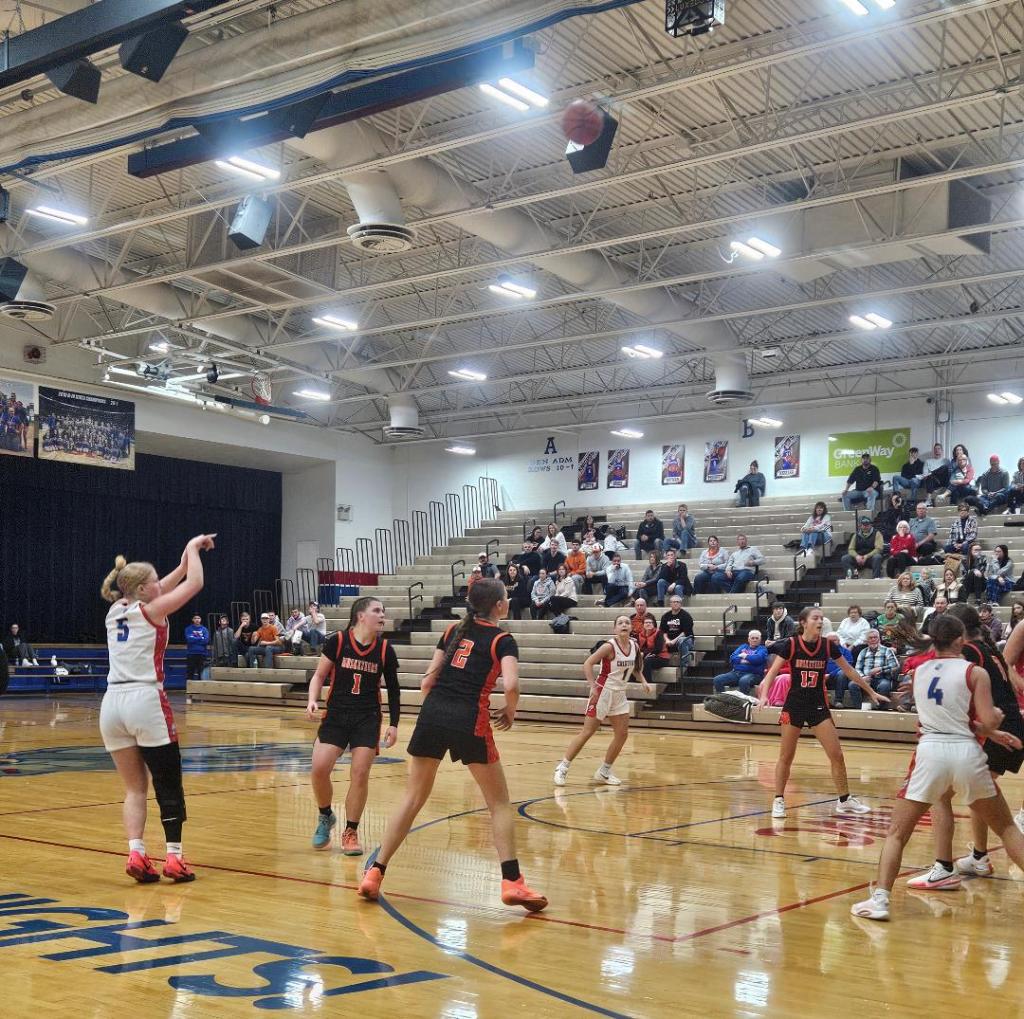 A basketball player wearing a white uniform and the number 5 attempts a shot while several opposing players in black uniforms watch closely. The gymnasium is filled with spectators and features bleachers in the background.