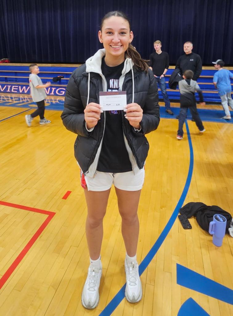 A female athlete stands in a gymnasium, smiling and holding a card, wearing a black jacket and white shorts, with other people in the background.