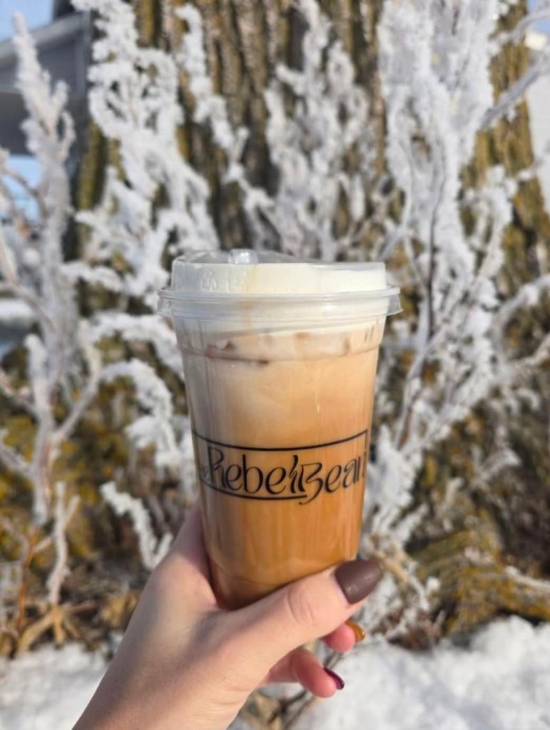 A hand holding a cup of iced coffee from The Rebel Bean, with frosty trees in the background.