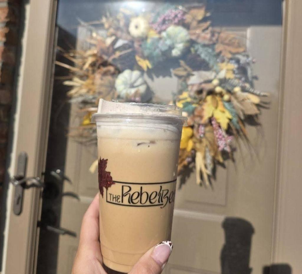 A hand holding a clear cup of iced coffee in front of a decorative autumn wreath on a door.