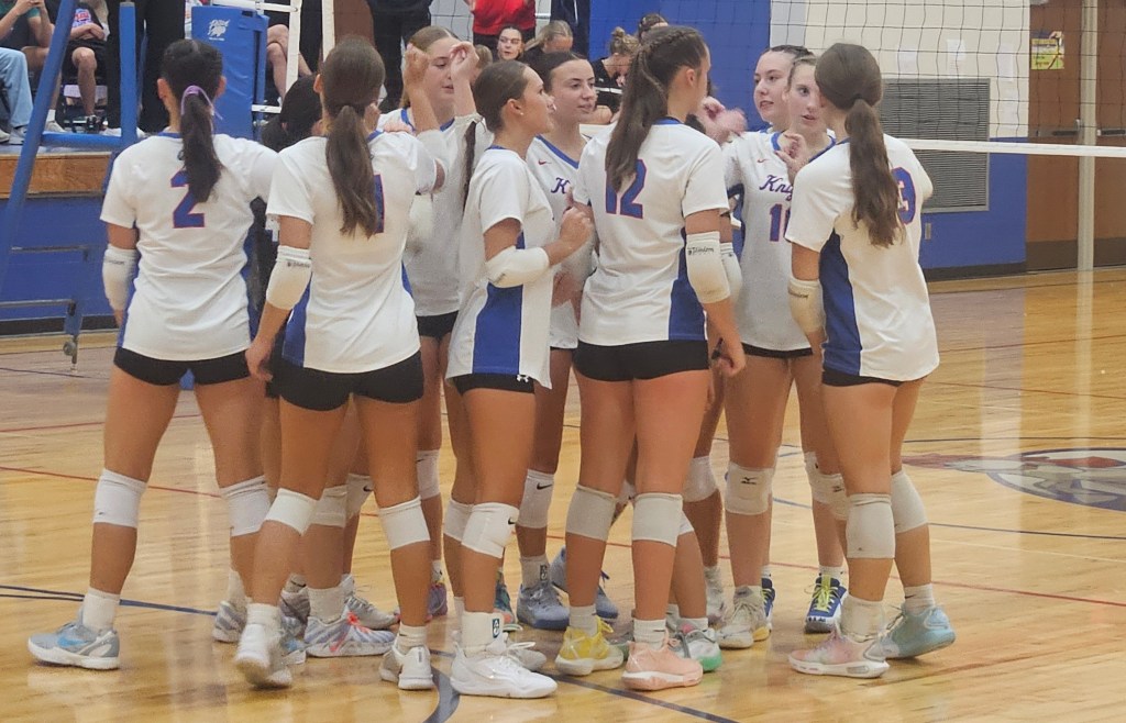 A group of young female volleyball players huddled together on a court, wearing matching white uniforms with blue accents, engaged in a team discussion.