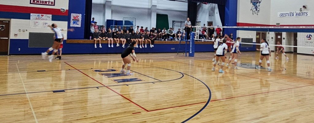 A volleyball match in a gymnasium, featuring players in action with a net in the center and spectators on the benches in the background.