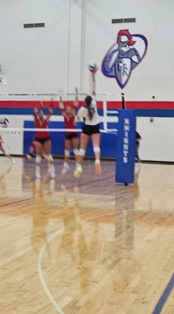 A high school volleyball game in action, featuring a player in mid-jump preparing to hit the ball over the net, while opposing players attempt to block the shot.