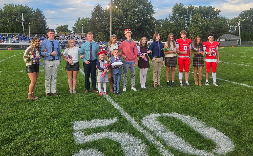 A group of students standing on a football field, dressed in formal attire for homecoming, with a football and bouquets in their hands. The backdrop includes spectators in the stands, trees, and a cloudy sky.