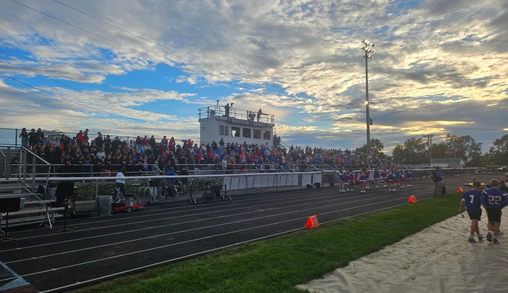 A packed grandstand at a high school football game, with fans in various attire and a vibrant sky in the background. Cheerleaders are on the field, and players walk nearby.