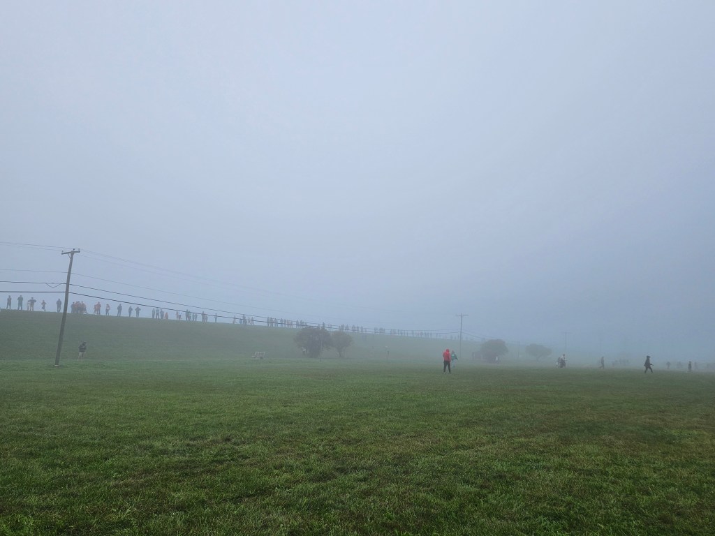 A foggy scene featuring a grassy field with indistinct figures scattered across it, some people walking while others are seen in the background on a hilltop, partially obscured by mist.
