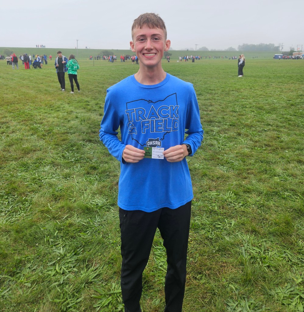 A smiling young athlete in a blue 'Track & Field' shirt holds an event card while standing on a grassy field with other people in the background.