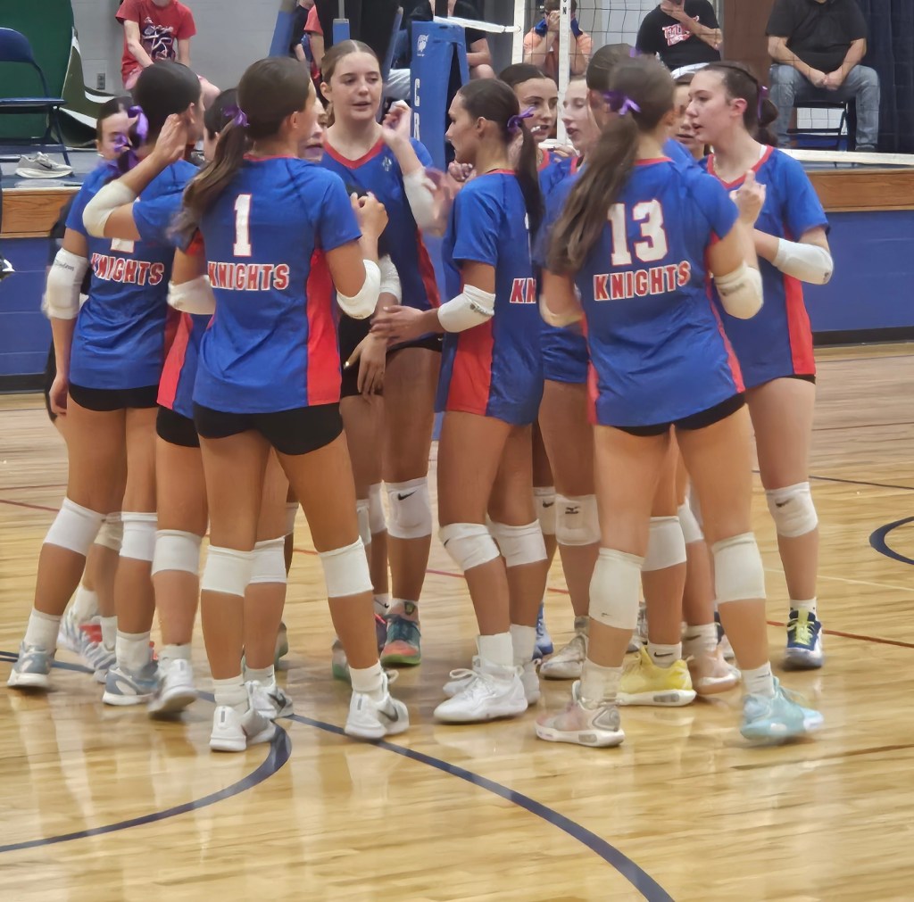 A group of female volleyball players wearing blue and red uniforms is gathered in a huddle, showing enthusiasm and teamwork on the court.