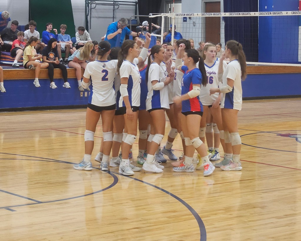 A volleyball team huddles together in a gymnasium, wearing matching uniforms while preparing for the game, with spectators seated in the background.
