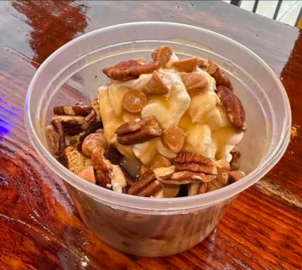 A clear plastic cup filled with a layered dessert featuring creamy toppings, chopped pecans, caramel drizzles, and crunchy bits.