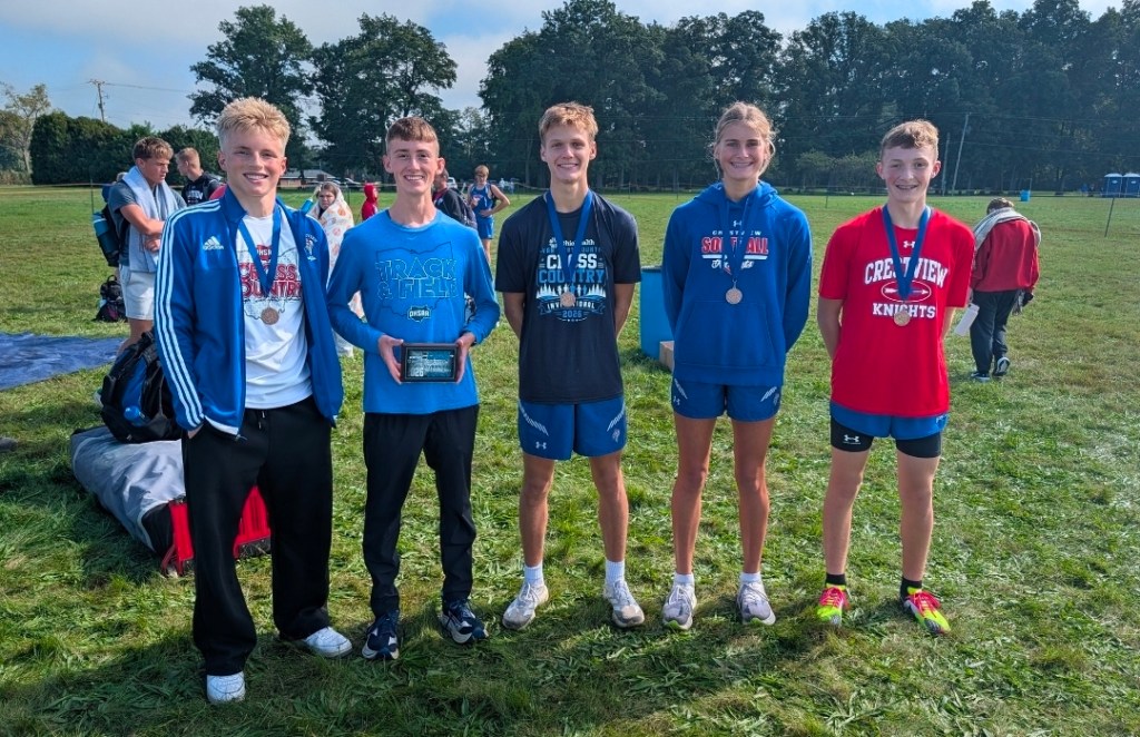 Five young athletes posing together in a grassy field after a track and field event, each wearing medals and team apparel, with a backdrop of spectators and trees.