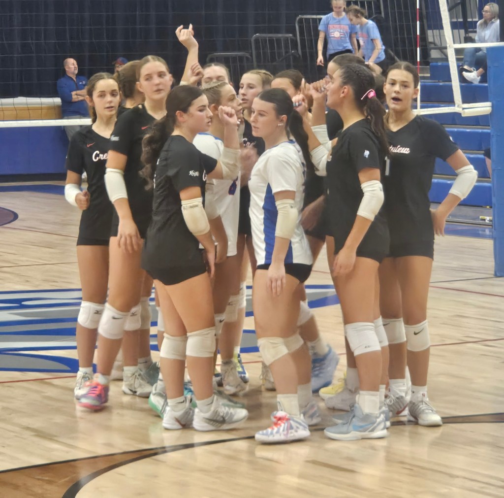 A volleyball team huddled together on the court, discussing strategy. The players are wearing black and white uniforms with knee pads, showing a mix of engagement and focus.