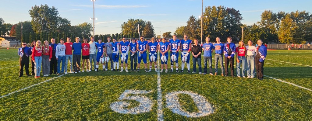 A group photo on a football field, featuring a team of players in blue uniforms standing in the center, surrounded by supporters wearing red and blue attire. The scene is set during sunset with trees in the background.