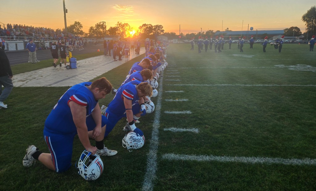 High school football team kneeling on the sidelines during sunset, with a crowd in the background and a marching band visible on the field.